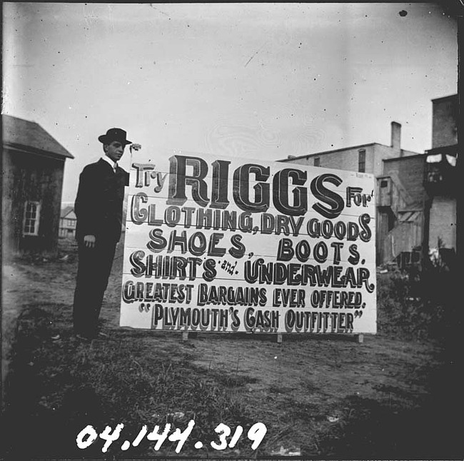 Man Holding up a Large Sign Advertising Riggs Store