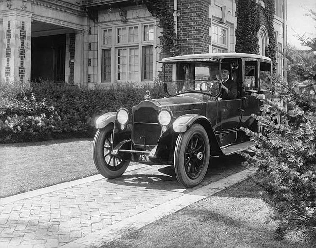 1918-1919 Packard limousine, parked in driveway with male chauffeur, next to large brick home