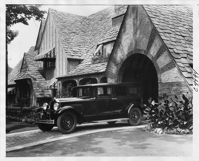 1930 Packard sedan limousine, three-quarter left side view, parked on drive at side of large brick home