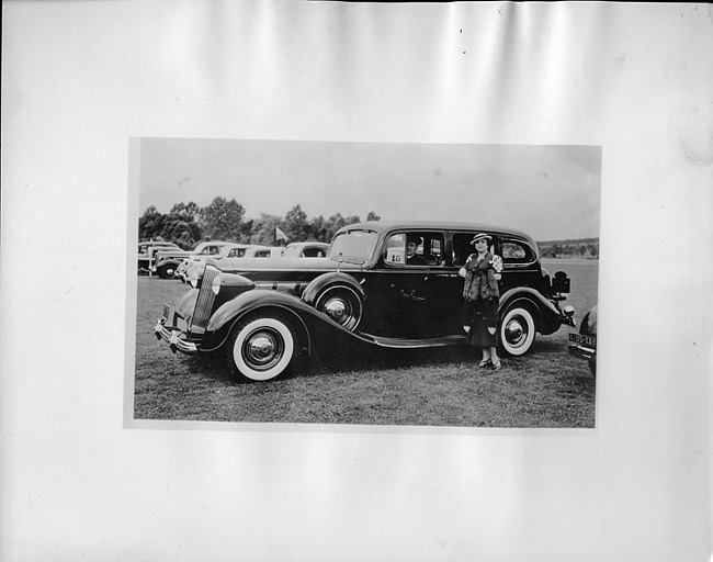 1937 Packard touring sedan with male chauffeur, female standing at passenger door