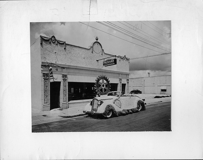 1937 Packard coupe roadster, top folded, parked on street in front of Packard dealership