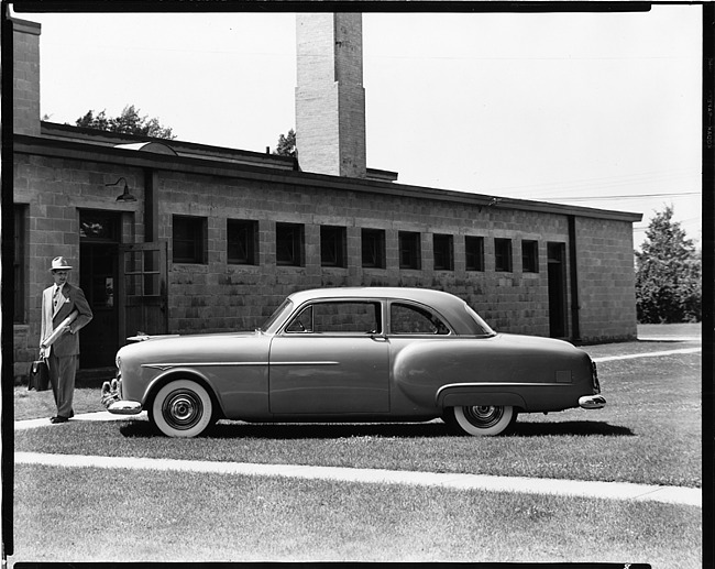 1951 Packard 200 sedan, parked on grass in front of building, man standing nearby