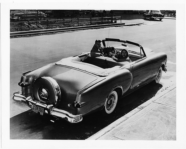 1953 Packard convertible, top folded, couple sitting in front seat, parked on street