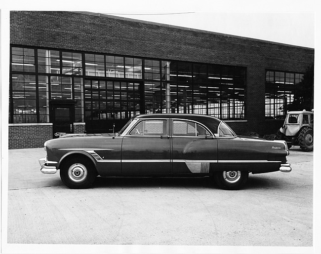 1953 Packard sedan, left side view, parked on drive, next to brick building