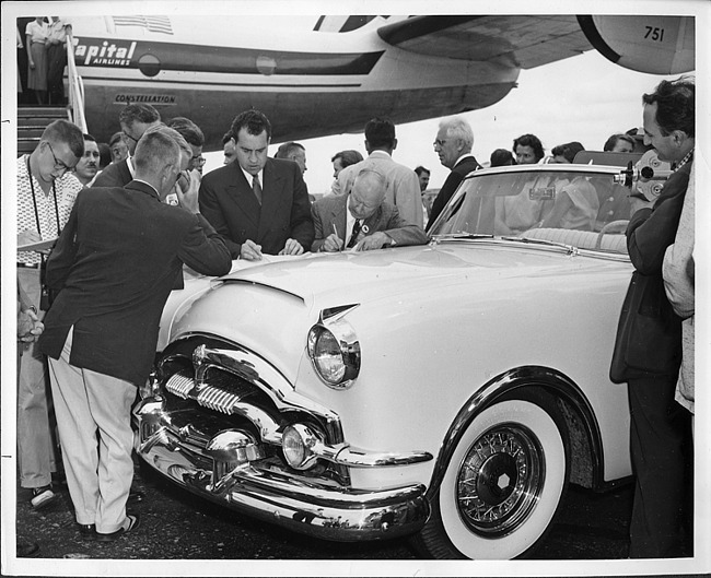 Vice-president Nixon at airport, looking over papers while leaning on a 1954 Packard Caribbean