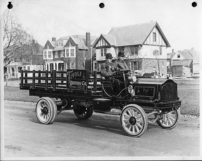 Early 1900s Packard truck, right side view, parked on residential street houses in background, two men in seat