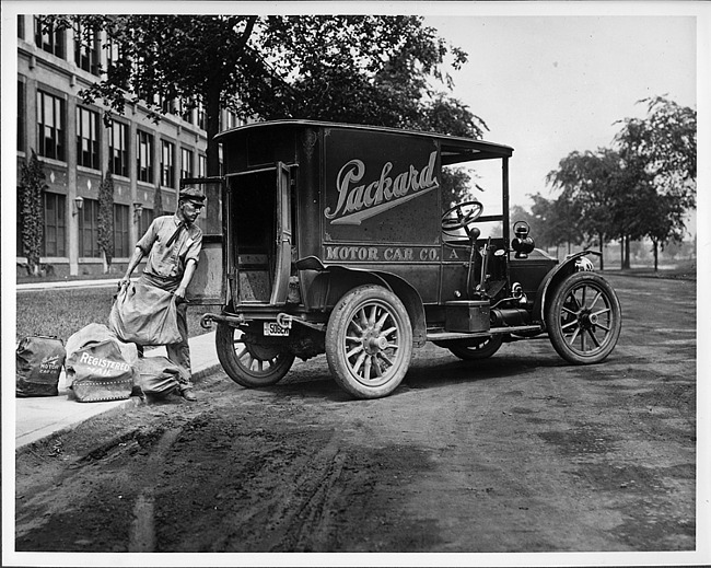 1910 Packard truck, parked on road, man loading mail into back of truck