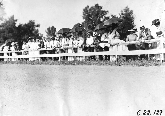 Group of men and women stand behind rail fence at a race track in Minnesota, at the 1909 Glidden Tour
