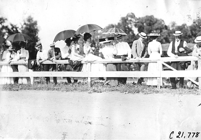 Group of men and women stand behind rail fence at a race track in Minnesota, at the 1909 Glidden Tour