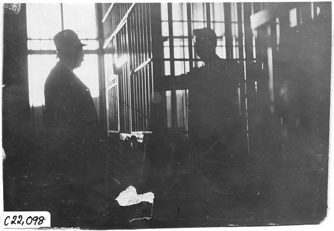 Man standing in jail cell police officer stands opposite him, at 1909 Glidden Tour