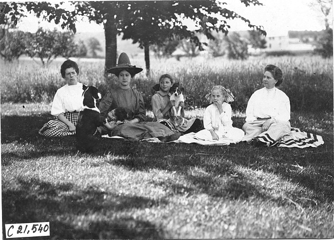 Women, girls and dogs sit on grass, at 1909 Glidden Tour