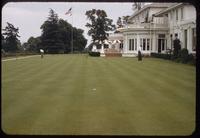 View of the practice green, with 2 golfers, in front of the clubhouse of the Los Angeles Country Club, 1954