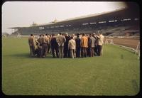 A group of men in a circle viewing turf on the inside track at Santa Anita Park racetrack, California, 1953, with the dirt track and the grandstand in the background, during a field day event